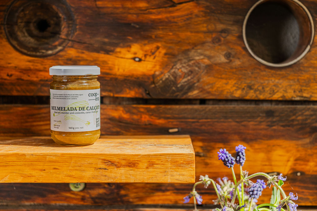 Pot de mel artesana sobre un bloc de fusta amb flors d'espígol, representant els productes de l'economia social i solidària del Penedès.